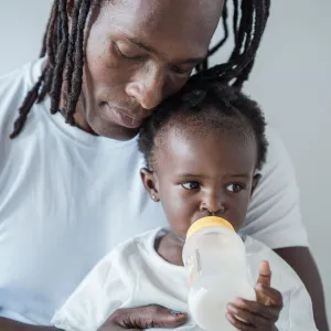 Image on the left of a young girl drinking milk out of a bottle while being held by her father. Photo by Sasha Kim.