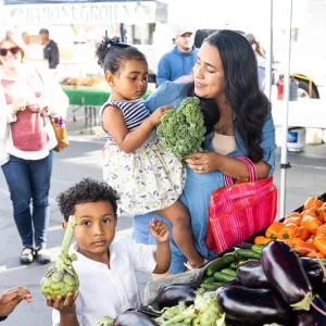 A mother holds her daughter in her arms while her son stands next to her holding fresh vegetables he selected at a farmers' market booth.