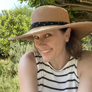 A woman in a hat and striped top smiles in a green garden.