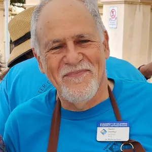 A smilingn man with a beard wearing a teal shirt and a dark blue apron.
