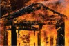 A wooden structure engulfed in intense orange flames, with charred beams and a roof that is about to collapse visible.